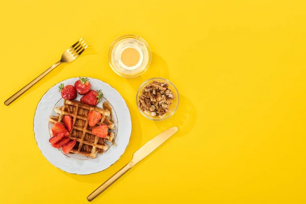 Сверху вид на подаваемый завтрак с вафлями, ягодами, медом и орехами возле столовых приборов на желтый — стоковое фото