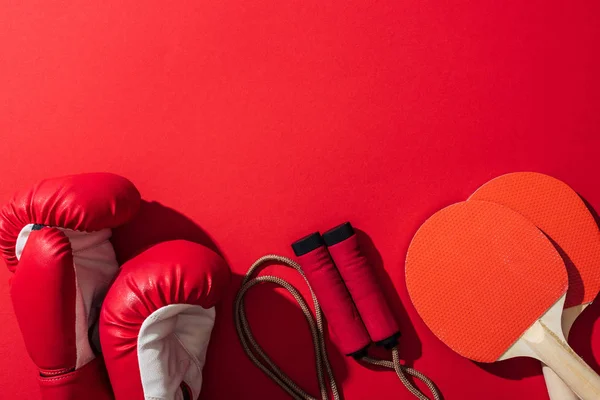 Вид сверху боксерских перчаток рядом с красными ракетками для пинг-понга и скакалка на красном — стоковое фото