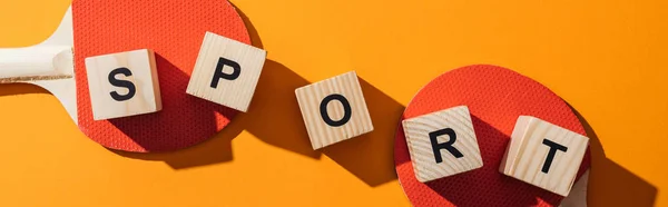 Panoramic shot of wooden cubes with sport lettering near table tennis rackets on yellow — Stock Photo