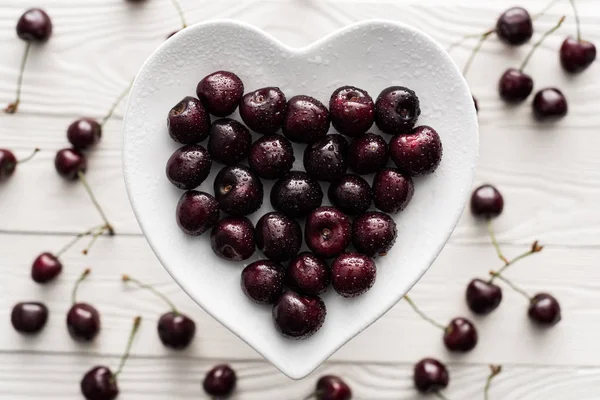 Вид сверху на свежие, сладкие и спелые вишни, покрытые капельками воды на тарелке в форме сердца — стоковое фото