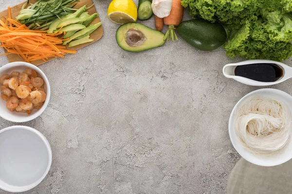 Top view of lemon, avocados, carrots, black pepper, shrimps, soy sauce, rice paper, garlic,noodles, sliced vegetables and lettuce on table — Stock Photo