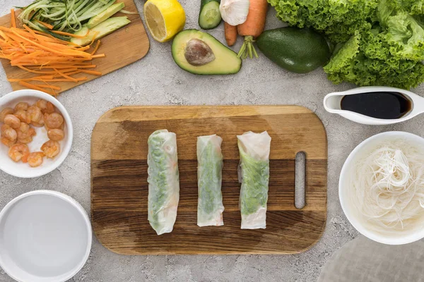 Vue du dessus des rouleaux de printemps sur la planche à découper parmi les ingrédients — Photo de stock