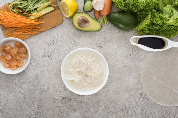 Top view of bowl with water and noodles, lemon, avocados, carrots, black pepper, shrimps, soy sauce, rice paper, garlic, sliced vegetables and lettuce on table — Stock Photo