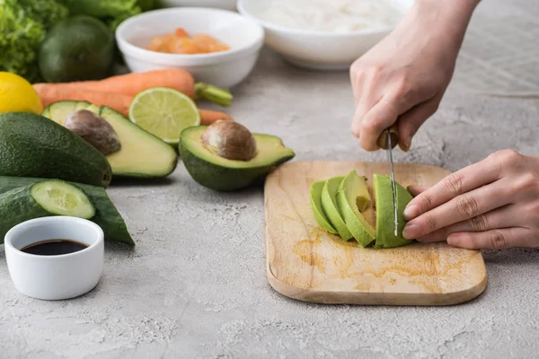 Vista ritagliata di donna taglio avocado con coltello sul tagliere tra gli ingredienti — Foto stock