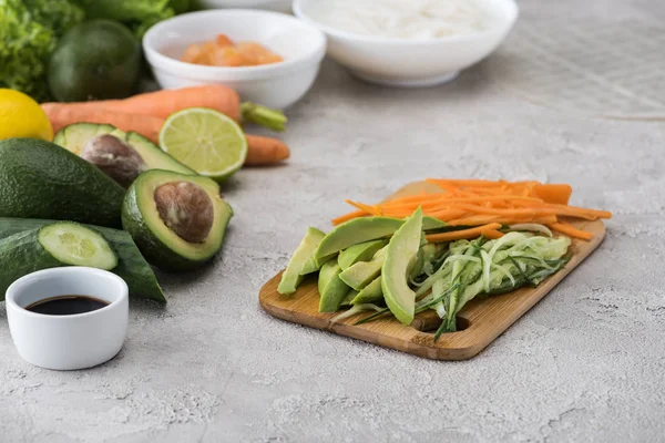 Cut avocado, carrot and cucumber on cutting board among raw ingredients — Stock Photo