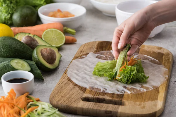 Cropped view of woman putting cut cucumber on carrot, lettuce and rice paper — Stock Photo