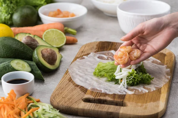 Vue recadrée d'une femme mettant des crevettes sur de la laitue, des nouilles et du papier de riz sur une planche à découper — Photo de stock