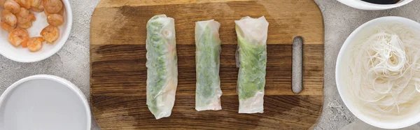 Panoramic shot of spring rolls on cutting board among ingredients — Stock Photo