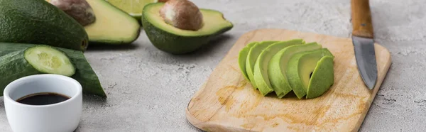 Панорамний знімок розрізаного авокадо на обробній дошці з ножем серед сирих інгредієнтів — стокове фото