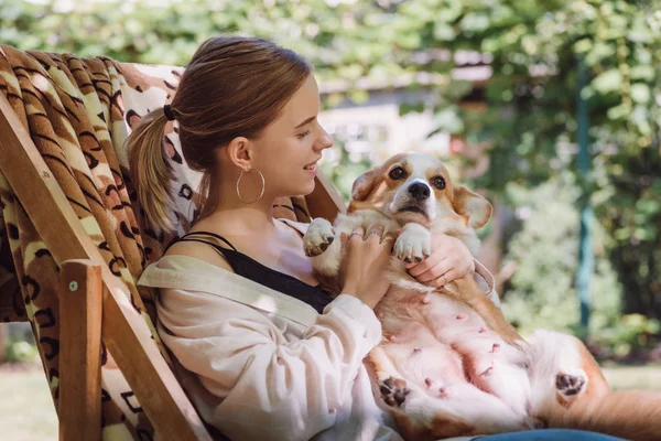 Cheerful blonde girl holding welsh corgi dog while sitting in deck chair in garden — Stock Photo