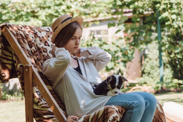 Chica rubia en sombrero de paja sosteniendo cachorro de rodillas mientras está sentado en la silla de cubierta en el jardín - foto de stock