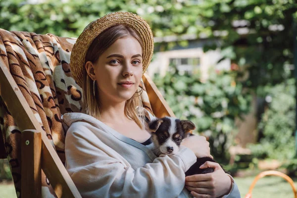 Happy blonde girl in straw hat holding puppy while sitting in deck chair in garden — Stock Photo