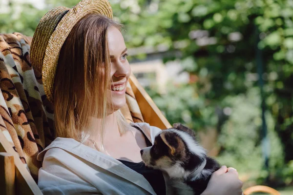 Счастливая блондинка в соломенной шляпе, держащая щенка корги, сидя в шезлонге в саду — стоковое фото