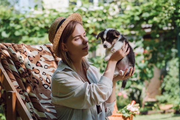 Rubia chica en paja sombrero celebración galés corgi cachorro mientras sentado en cubierta silla en jardín - foto de stock