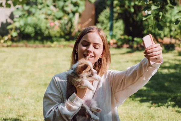 Happy blonde girl taking selfie with cute Welsh corgi puppy in garden — стоковое фото