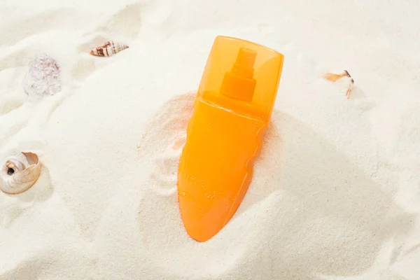 Оранжевая бутылка солнцезащитного крема в песке с ракушками — стоковое фото