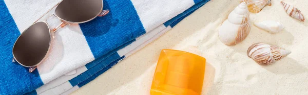 Botella naranja de protector solar en la arena con conchas marinas, toalla a rayas y gafas de sol, tiro panorámico - foto de stock