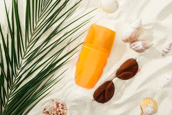 Orange bottle of sunscreen on sand with seashells, green palm leaf and stylish sunglasses — Stock Photo