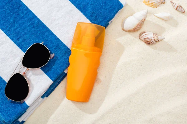 Помаранчева пляшка сонцезахисного крему на піску з черепашками, смугастим рушником та сонцезахисними окулярами — стокове фото