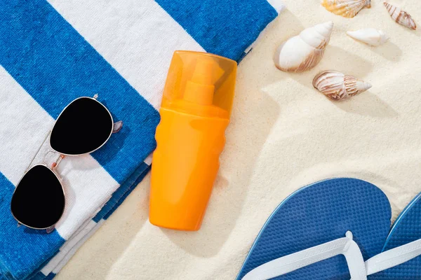 Botella naranja de protector solar en arena cerca de toalla rayada, chanclas azules, gafas de sol y conchas marinas - foto de stock