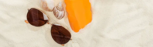 Botella naranja de protector solar en la arena cerca de conchas marinas y gafas de sol, tiro panorámico - foto de stock