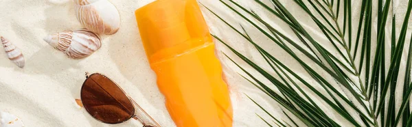 Bouteille orange de crème solaire sur sable avec coquillages, feuille de palme verte et lunettes de soleil élégantes, vue panoramique — Photo de stock
