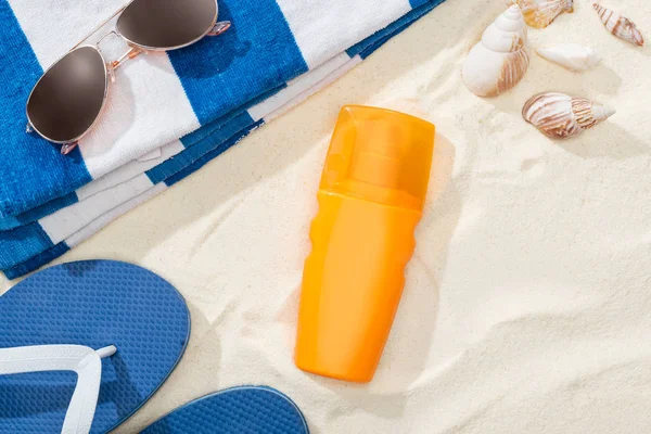Bottiglia di crema solare sulla sabbia vicino all'asciugamano a strisce, infradito blu, occhiali da sole e conchiglie — Foto stock
