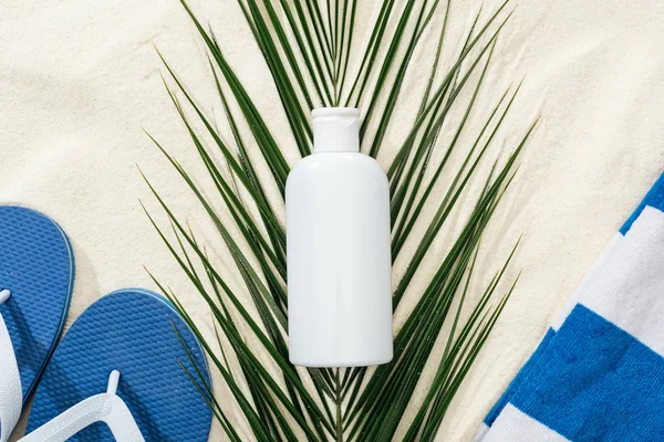 Білий сонцезахисний лосьйон на зеленому пальмовому листі на піску з блакитними фліпками та рушником — стокове фото
