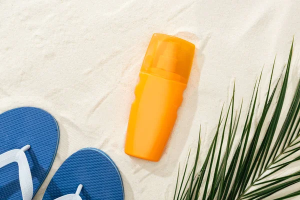 Protetor solar laranja perto de folha de palma verde na areia com chinelos azuis — Fotografia de Stock