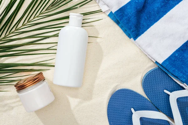 Loción de protector solar blanco y crema cerca de hoja de palma verde en la arena con chanclas azules y toalla - foto de stock