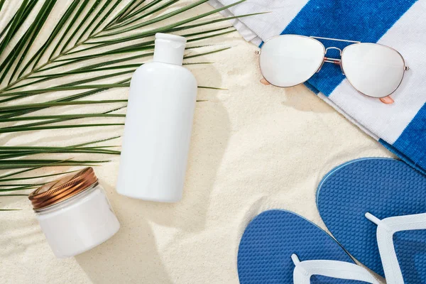 Lotion solaire blanche et crème près de la feuille de palme verte sur sable avec tongs bleues, lunettes de soleil et serviette — Photo de stock