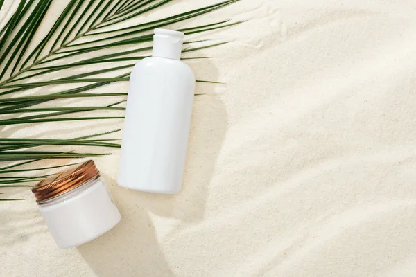 White sunscreen lotion and cream near green palm leaf on sand — Stock Photo