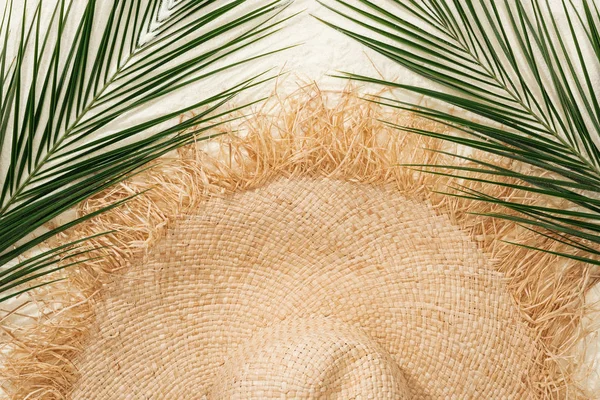 Vista superior de sombrero de paja elegante en arena dorada con hojas de palma verde - foto de stock