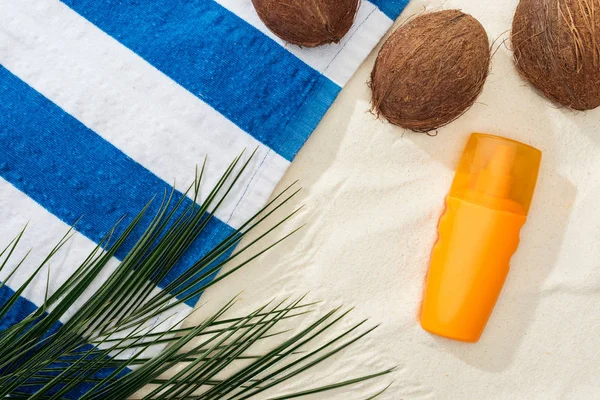 Vista superior de hojas de palma, cocos y toalla rayada cerca de protector solar naranja en arena - foto de stock