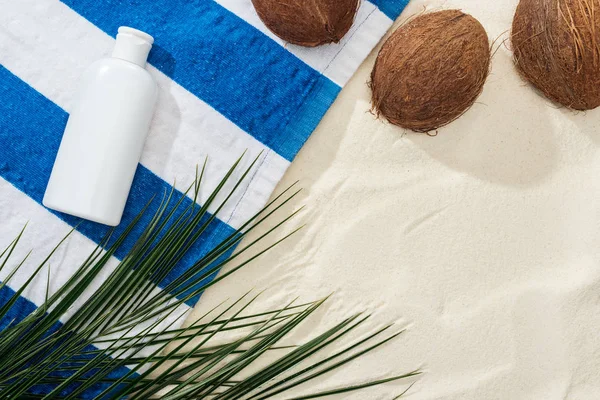 Vista superior de folhas de palmeira, cocos e toalha listrada com protetor solar branco na areia — Fotografia de Stock