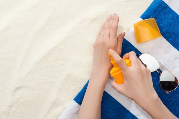 Vista recortada de la mujer que aplica protector solar en las manos cerca de la toalla rayada y gafas de sol en la arena - foto de stock