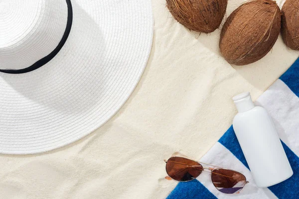Vista superior de gafas de sol, toalla rayada, cocos y sombrero de paja blanco cerca de loción blanca en la arena - foto de stock