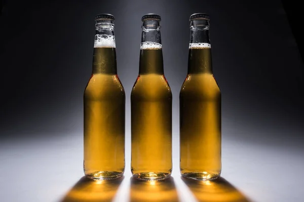 Três garrafas de vidro com cerveja no fundo escuro com luz traseira — Fotografia de Stock