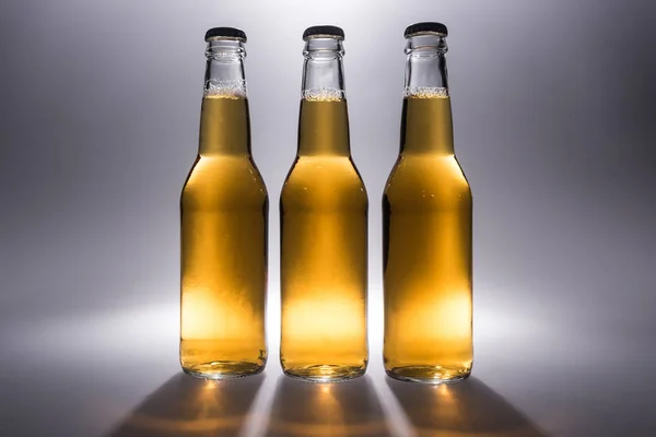 Tres botellas de vidrio con cerveza sobre fondo gris con luz de fondo - foto de stock