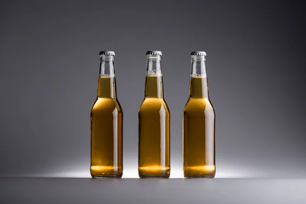 Três garrafas de vidro com cerveja em linha no fundo cinza — Fotografia de Stock