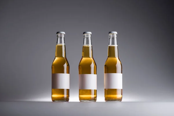 Tre bottiglie di vetro con birra in fila con etichette bianche su sfondo grigio — Foto stock