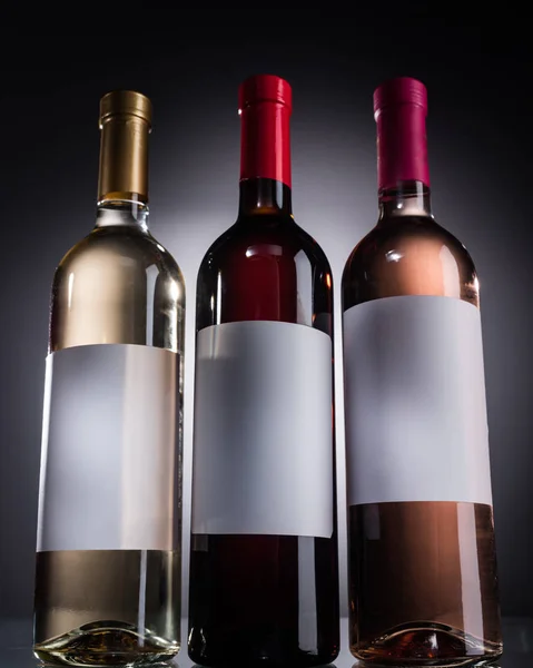 Vista de ángulo bajo de botellas con vino blanco, rojo y rosa y etiquetas en blanco sobre fondo oscuro con luz de fondo - foto de stock
