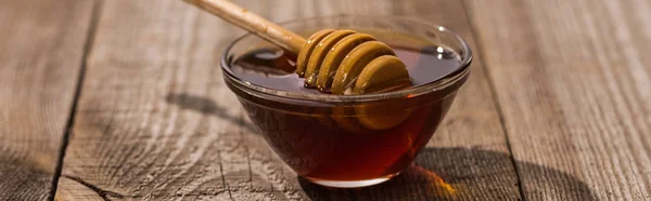 Panoramic shot of glass bowl with honey and honey dipper on wooden table in sunlight — Stock Photo