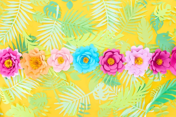 Disposición plana con flores multicolores de corte de papel en fila sobre hojas tropicales sobre fondo amarillo brillante - foto de stock