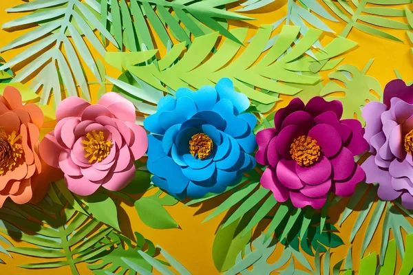 Deitado plano com papel cortar flores multicoloridas em folhas tropicais no fundo amarelo brilhante — Fotografia de Stock