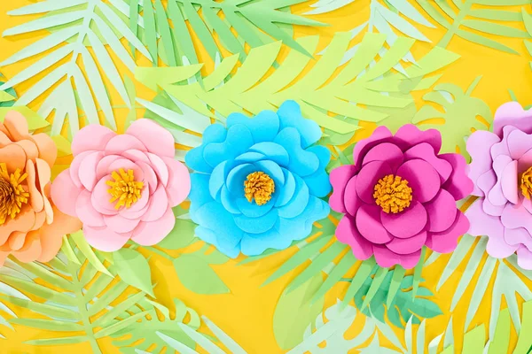 Disposición plana con flores multicolores de corte de papel sobre hojas tropicales sobre fondo amarillo brillante - foto de stock