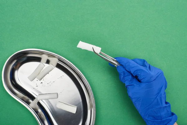 Cropped view of dentist holding tweezers with cotton pad near metallic plate on green — Stock Photo