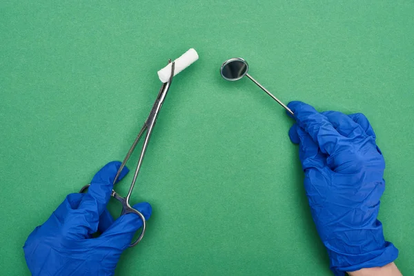 Vue recadrée du dentiste en gants de latex bleu tenant des ciseaux avec tampon de coton et miroir médical sur vert — Photo de stock