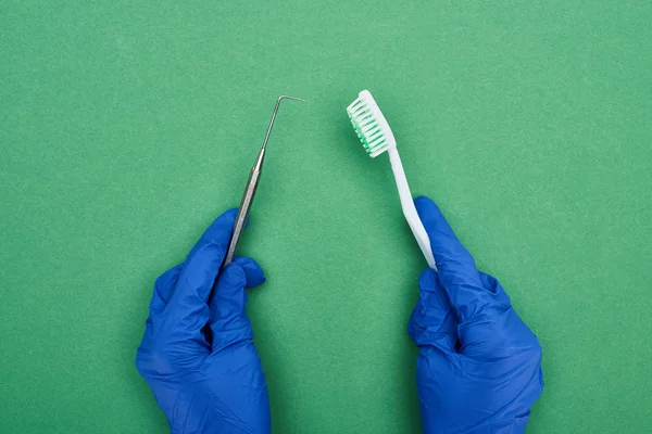 Cropped view of dentist holding dental scraper and toothbrush on green — Stock Photo