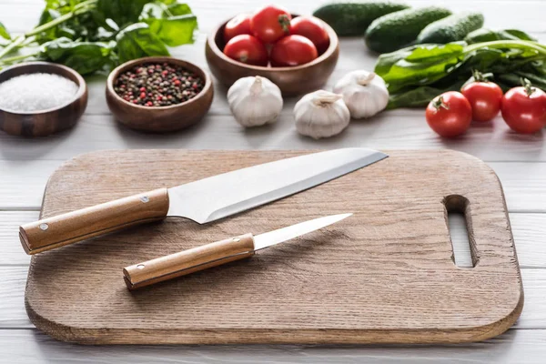 Foco seletivo de tábua de corte, tomate cereja, vegetação, garlics, facas, pepinos, sal e especiarias — Fotografia de Stock